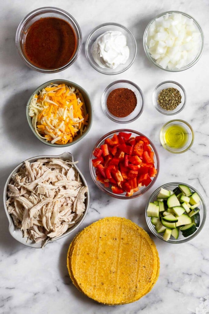 White marble counter with a bowl of diced white onion, two small bowls of different spices, a bowl of olive oil, a bowl of diced red bell pepper, a bowl of diced zucchini, a pile of corn tortillas, a bowl of shredded chicken, a bowl of shredded cheese, a bowl of greek yogurt, and a bowl of red enchilada sauce.