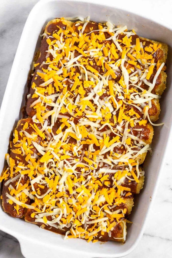 Casserole dish filled with chicken enchiladas covered in red sauce and shredded cheese before they are baked.