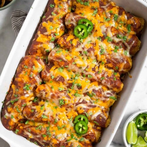 Healthy chicken enchilada with red sauce and cheese in a large casserole dish. They are garnished with sliced jalapeños and cilantro. Around the dish is a bowl of lime wedges, a bowl of cilantro, and a bowl of tortilla chips.