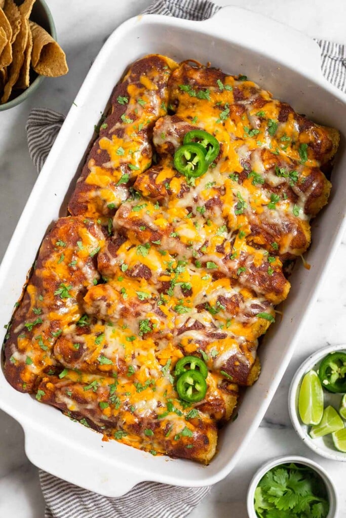 Healthy chicken enchilada with red sauce and cheese in a large casserole dish. They are garnished with sliced jalapeños and cilantro. Around the dish is a bowl of lime wedges, a bowl of cilantro, and a bowl of tortilla chips.