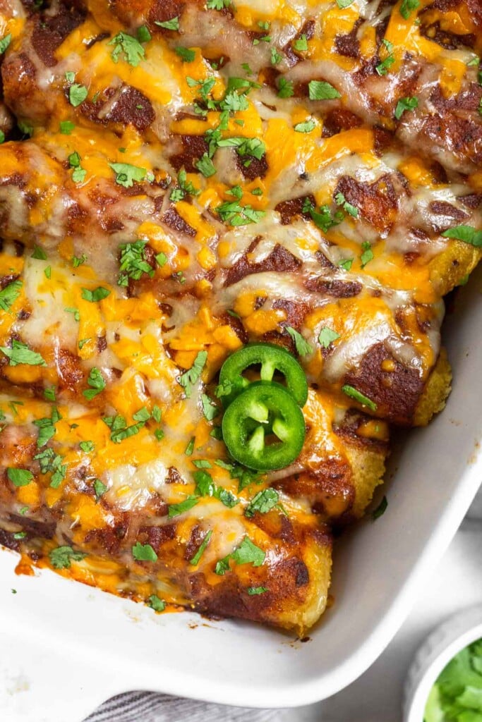 Overhead shot of healthy red chicken enchiladas in a casserole dish. They are topped with cilantro and sliced jalapeños.