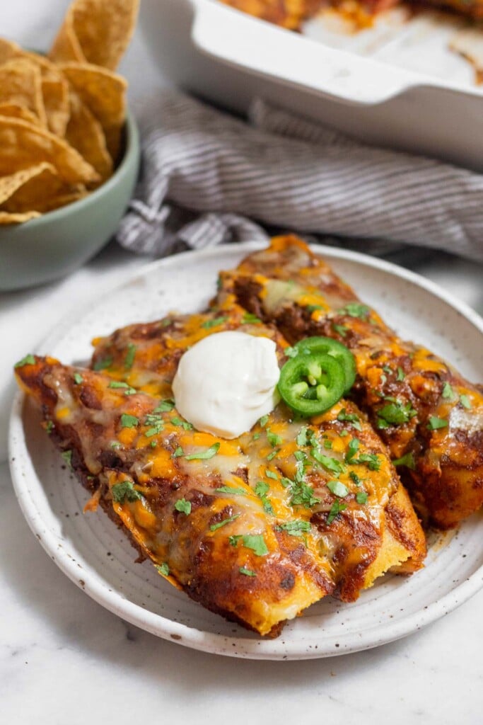 Three chicken enchiladas with red sauce and cheese on a plate. They are topped with cilantro, sliced jalapeños, and sour cream. Behind them is a bowl of tortilla chips and a casserole dish.