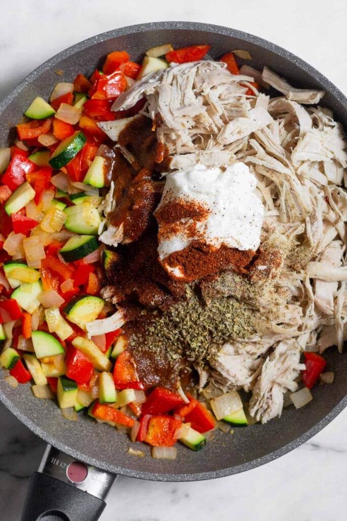 Skillet filled with sautéed onion, pepper, and zucchini. Piled on top of the veggies is shredded chicken, greek yogurt, enchilada sauce, and spices.