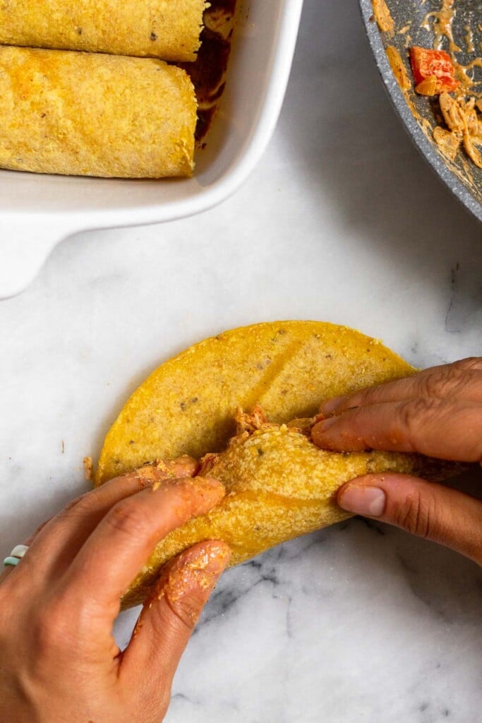 Chicken enchilada being rolled up. Next to it is a dish filled with enchiladas.
