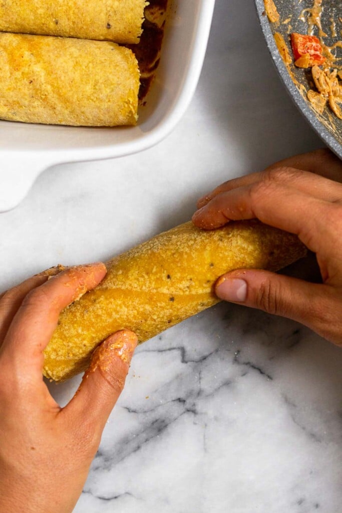 Chicken enchilada being rolled up. Next to it is a dish filled with enchiladas.