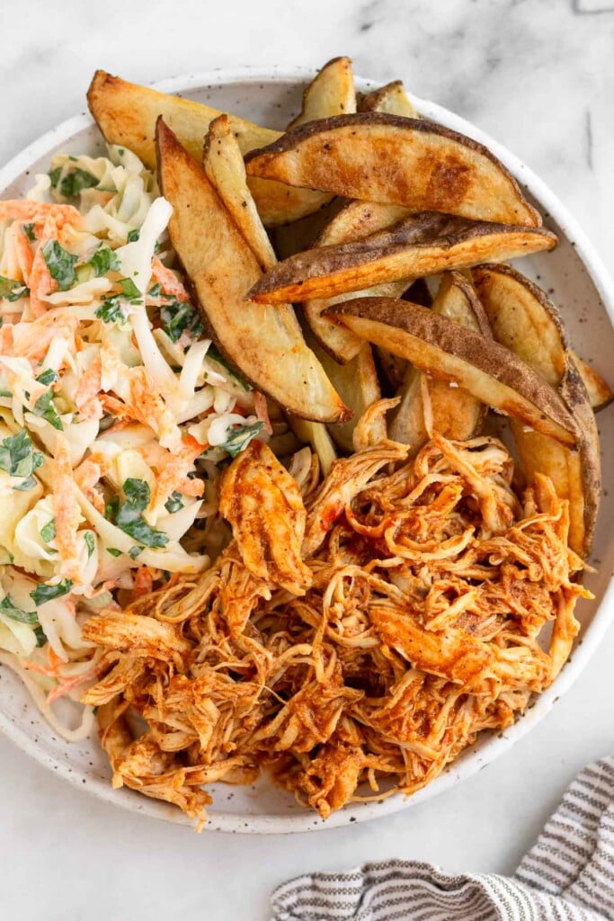 A white plate of pulled BBQ chicken, coleslaw, and potato wedges. Next to the plate is a striped kitchen towel.