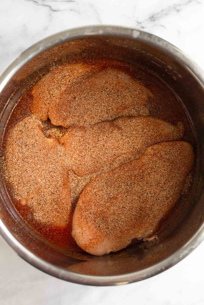 Large pot filled with barbecue sauce and raw chicken breasts with spices on them.
