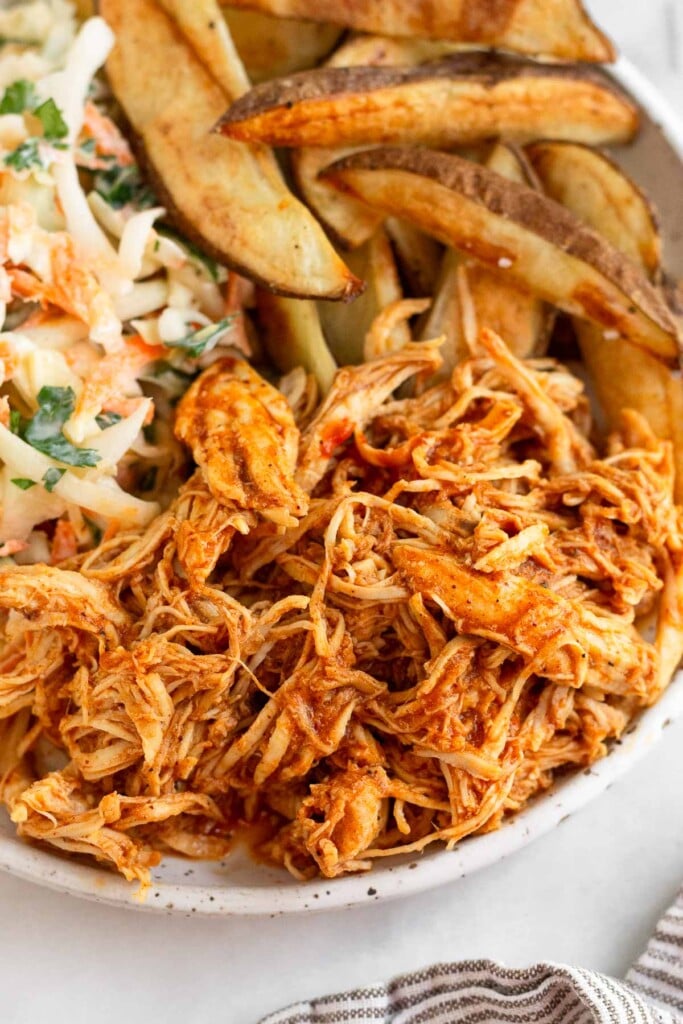 A close up on a white plate with shredded bbq chicken on it. Behind it is a pile of potato wedges and some coleslaw.