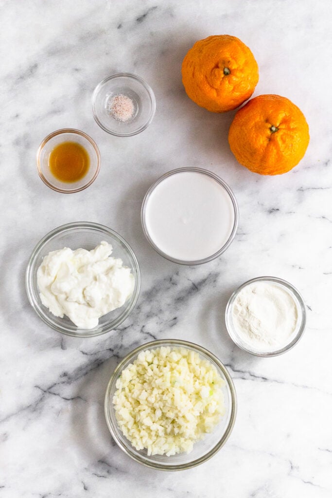 White counter top with 2 oranges, bowl of coconut milk, bowl of collagen, bowl of frozen cauliflower rice, bowl of greek yogurt, bowl of vanilla extract, and bowl of salt.