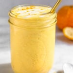 Orange creamsicle smoothie in a large glass with a straw in it. It is topped with some coconut milk and orange zest. Around it is some orange segments, a spoon, and a whole orange.