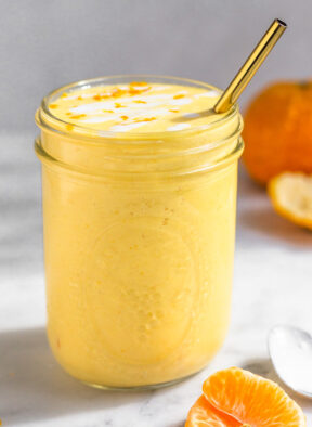 Orange creamsicle smoothie in a large glass with a straw in it. It is topped with some coconut milk and orange zest. Around it is some orange segments, a spoon, and a whole orange.