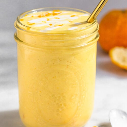 Orange creamsicle smoothie in a large glass with a straw in it. It is topped with some coconut milk and orange zest. Around it is some orange segments, a spoon, and a whole orange.