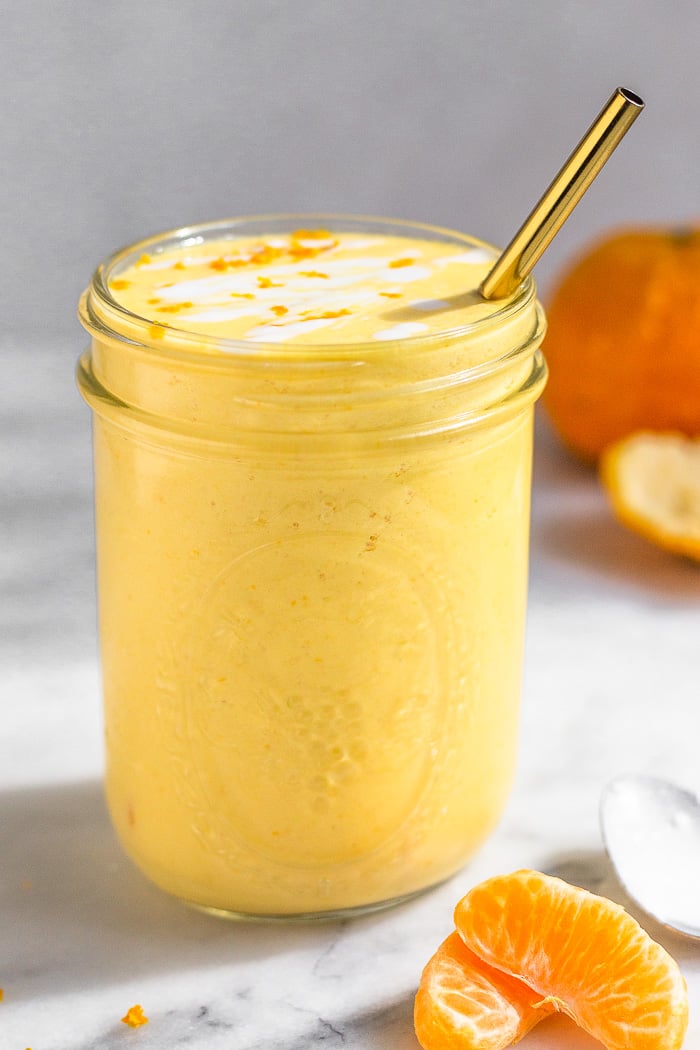 Orange creamsicle smoothie in a large glass with a straw in it. It is topped with some coconut milk and orange zest. Around it is some orange segments, a spoon, and a whole orange.