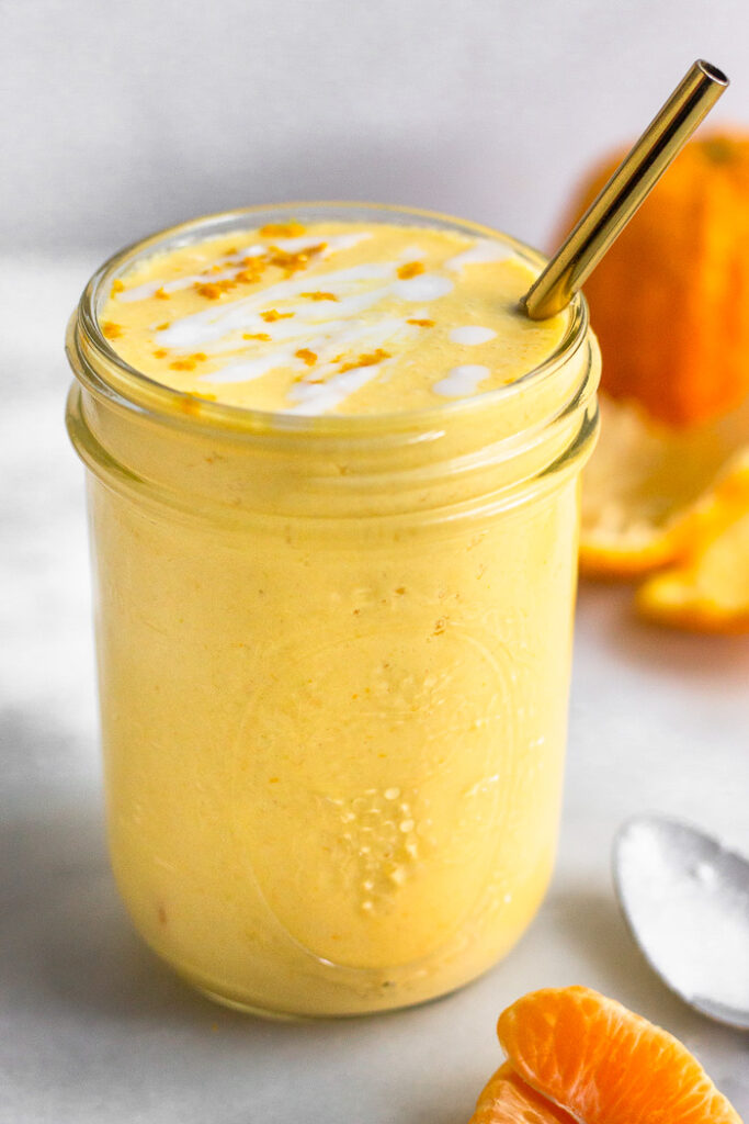 Creamsicle smoothie in a glass topped with coconut milk and orange zest with a straw in it. Around it is more oranges and a spoon with coconut milk on it.