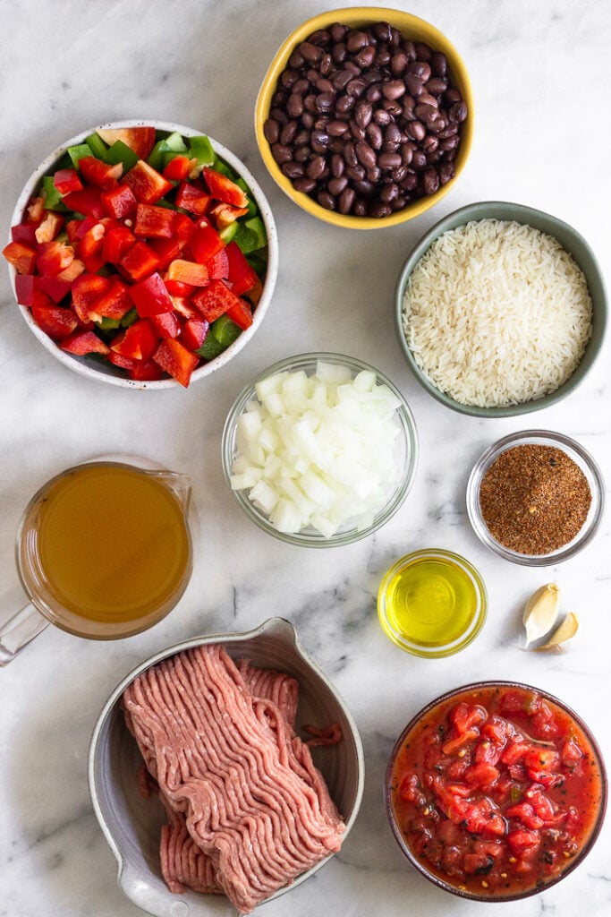 White counter top with a bowl of black beans, bowl of white rice, bowl of taco seasoning, 2 garlic cloves, bowl of diced canned tomatoes, bowl of raw ground turkey, bowl of olive oil, bowl of diced onion, jar of broth, and bowl of diced bell peppers.
