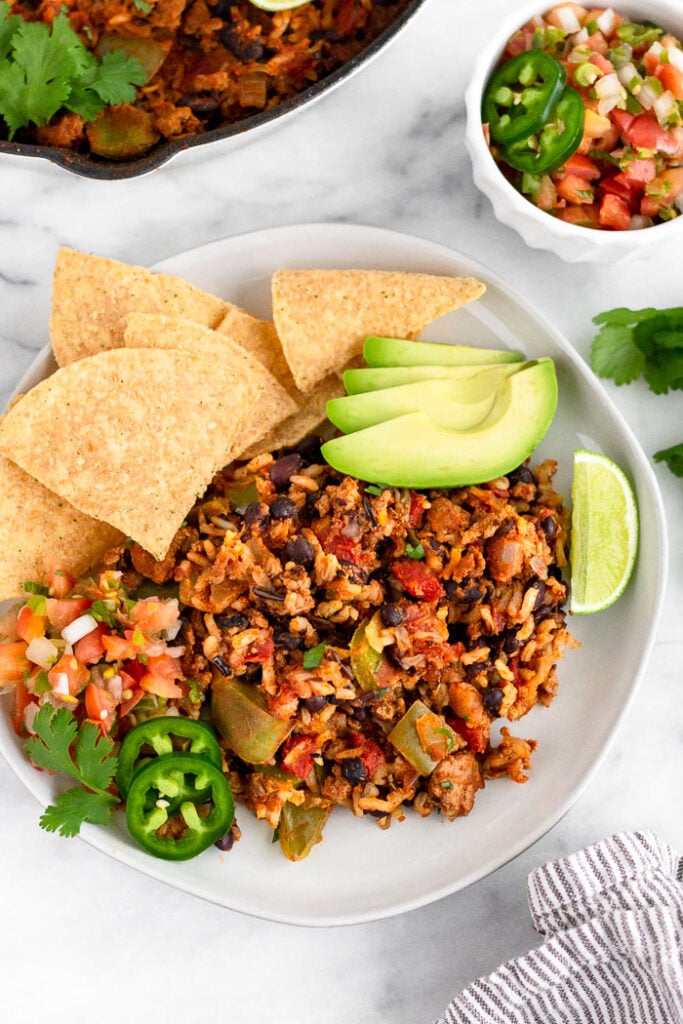 Plate of ground turkey taco skillet with some chips, sliced avocado, lime wedge, jalapeños, salsa, and cilantro. Next to is a bowl of most salsa and a pan of more of the taco skillet.
