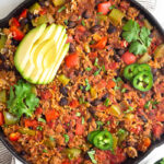 Turkey taco skillet in a large cast iron topped with avocado, jalapeño, and cilantro. Around it is a bowl of chips, bowl of cilantro, and a plate of lime wedges.
