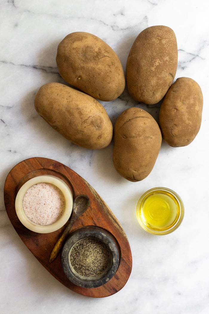 Overhead shot of 5 russet potatoes, bowl of olive oil, and a container of salt and pepper.