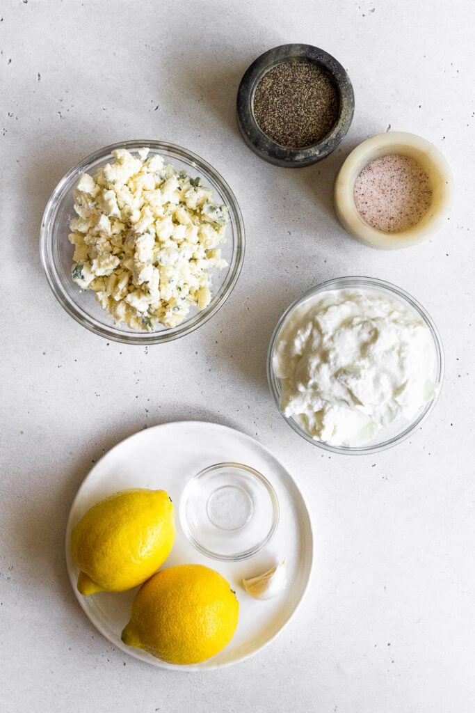 Overhead shot of small containers of salt and pepper, bowl of greek yogurt, plate of two lemons and a small bowl of vinegar, and bowl of blue cheese crumbles.