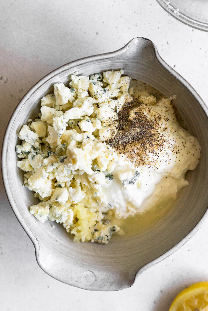 Overhead shot of a bowl with yogurt, minced garlic, blue cheese crumbles, lemon juice, and salt and pepper.