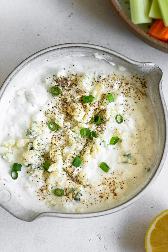 Overhead shot of greek yogurt blue cheese dressing topped with green onions and pepper. Next to it is half a lemon and plate of carrots and celery.