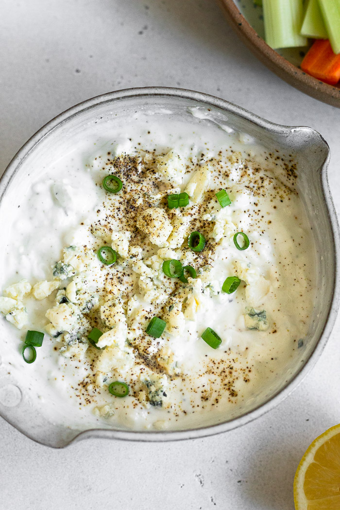 Overhead shot of greek yogurt blue cheese dressing topped with green onions and pepper. Next to it is half a lemon and plate of carrots and celery.