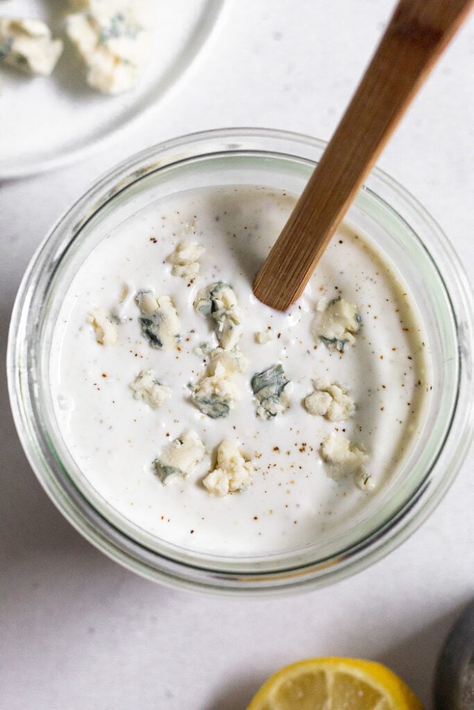 Jar of yogurt blue cheese dressing. There is a spatula in the jar and next to it is a plate of blue cheese and half a lemon.