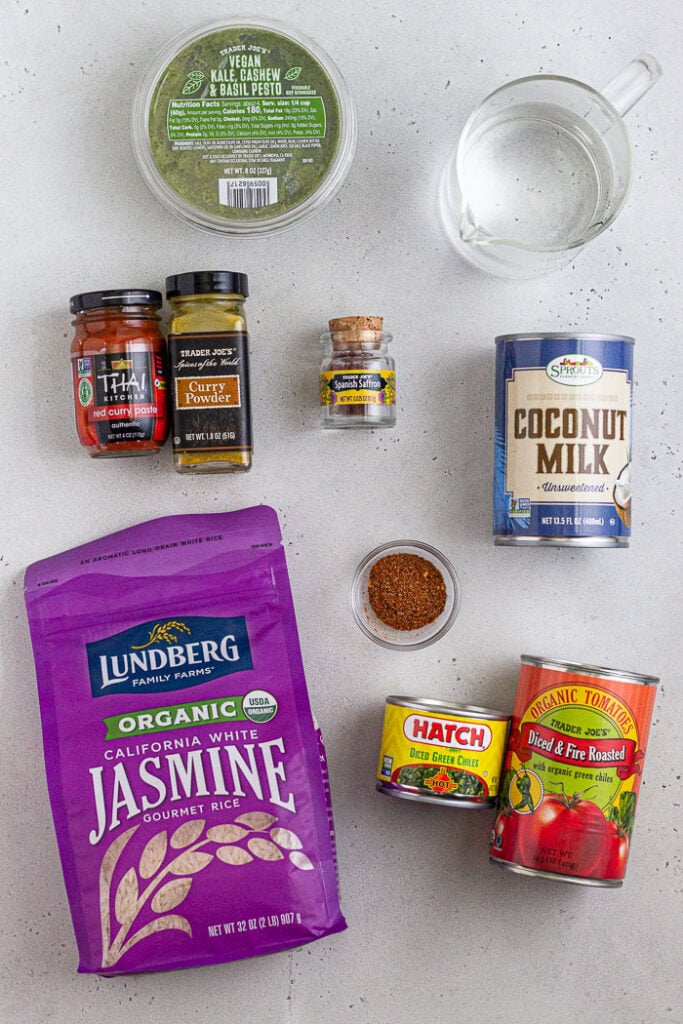 White counter with a container of pesto, cup of water, can of coconut milk, can of fire roasted tomatoes, can of green chilis, bowl of taco seasoning, bag of jasmine rice, jar of saffron, jar of curry powder, and a jar of curry paste.