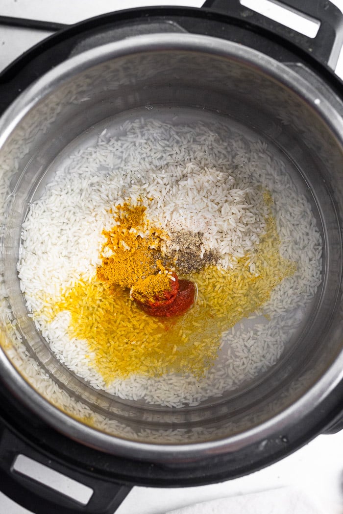 Instant pot bowl with uncooked white rice, water, curry paste, curry powder, and salt and pepper in it.