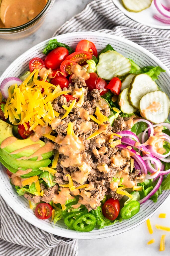 High protein salad with ground beef, cheese, avocado, tomatoes, pickles, onions, and special sauce dressing in a bowl. The bowl is on a towel and around it is more ingredients and a jar of more dressing.