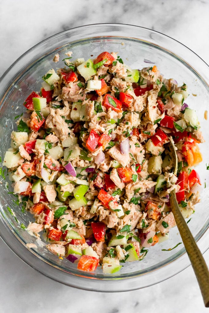 Tuna salad in a large glass bowl with veggies, fruit, and a spoon in it.