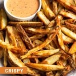 Air fryer french fries on a plate sprinkled with salt. There is also a small dish of orange sauce on the plate.