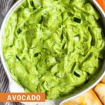 Avocado tzatziki dip in a bowl. Around it is pita chips, cherry tomatoes, carrot sticks, and sliced cucumber.