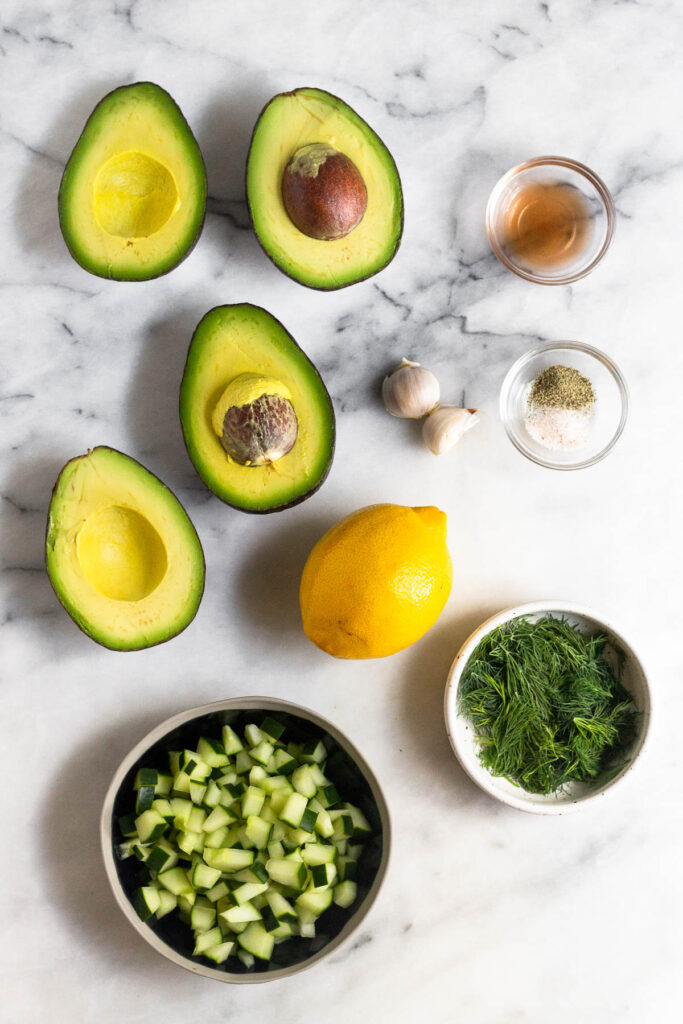 Overhead shot of 4 halves of avocado, small bowl of red wine vinegar, small bowl of salt and pepper, bowl of dill, whole lemon, and a bowl of diced cucumber.