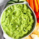 Avocado tzatziki dip in a bowl. Around it is pita chips, cherry tomatoes, carrot sticks, and sliced cucumber.