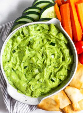 Avocado tzatziki dip in a bowl. Around it is pita chips, cherry tomatoes, carrot sticks, and sliced cucumber.