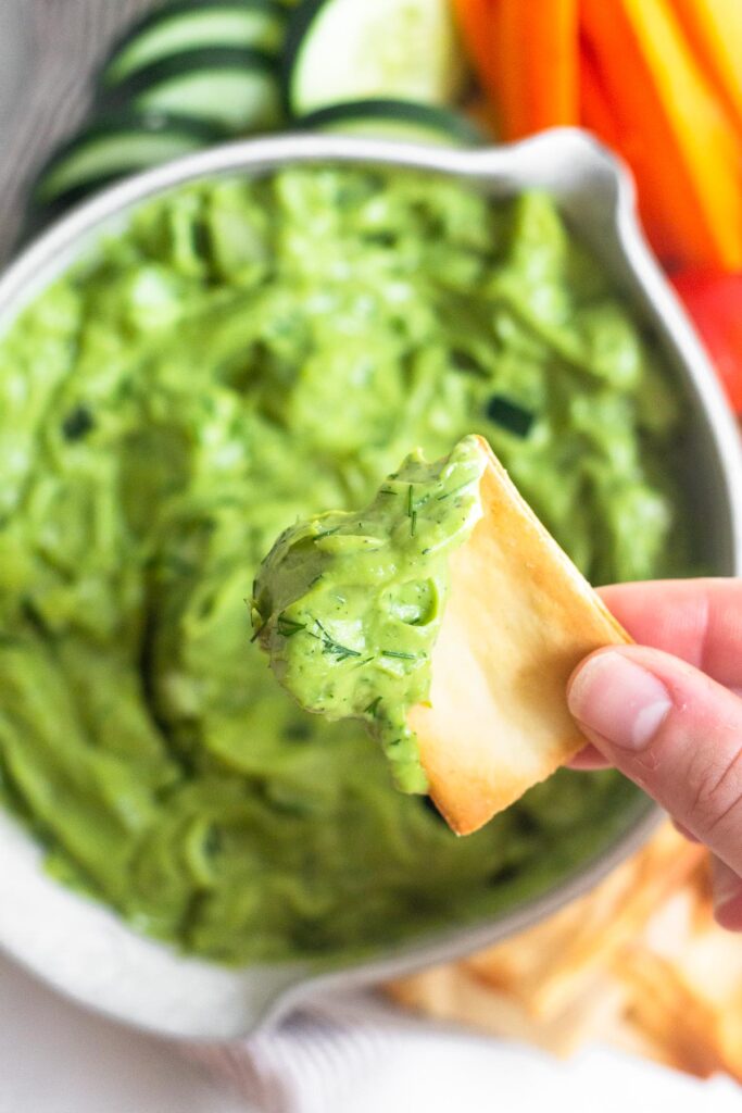Overhead shot of a pita chip with avocado tzatziki on it. Underneath is a bowl with more dip and some veggies and chips around it.