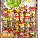 Grilled greek chicken kabobs on a baking sheet with a bowl of avocado tzatziki next to them.