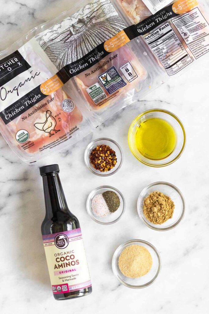 Overhead shot of a package of raw chicken thighs, bowl of olive oil, bowl of ground ginger, bowl of garlic powder, bowl of salt and pepper, jar of coconut aminos, and a bowl of red pepper flakes.