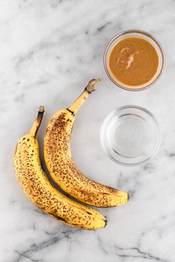 White counter with 2 spotted bananas, bowl of peanut butter, and a bowl of coconut oil.