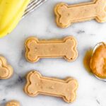 Overhead shot of peanut butter banana dog treats shaped as bones with a spoonful of peanut butter and bananas next to them.