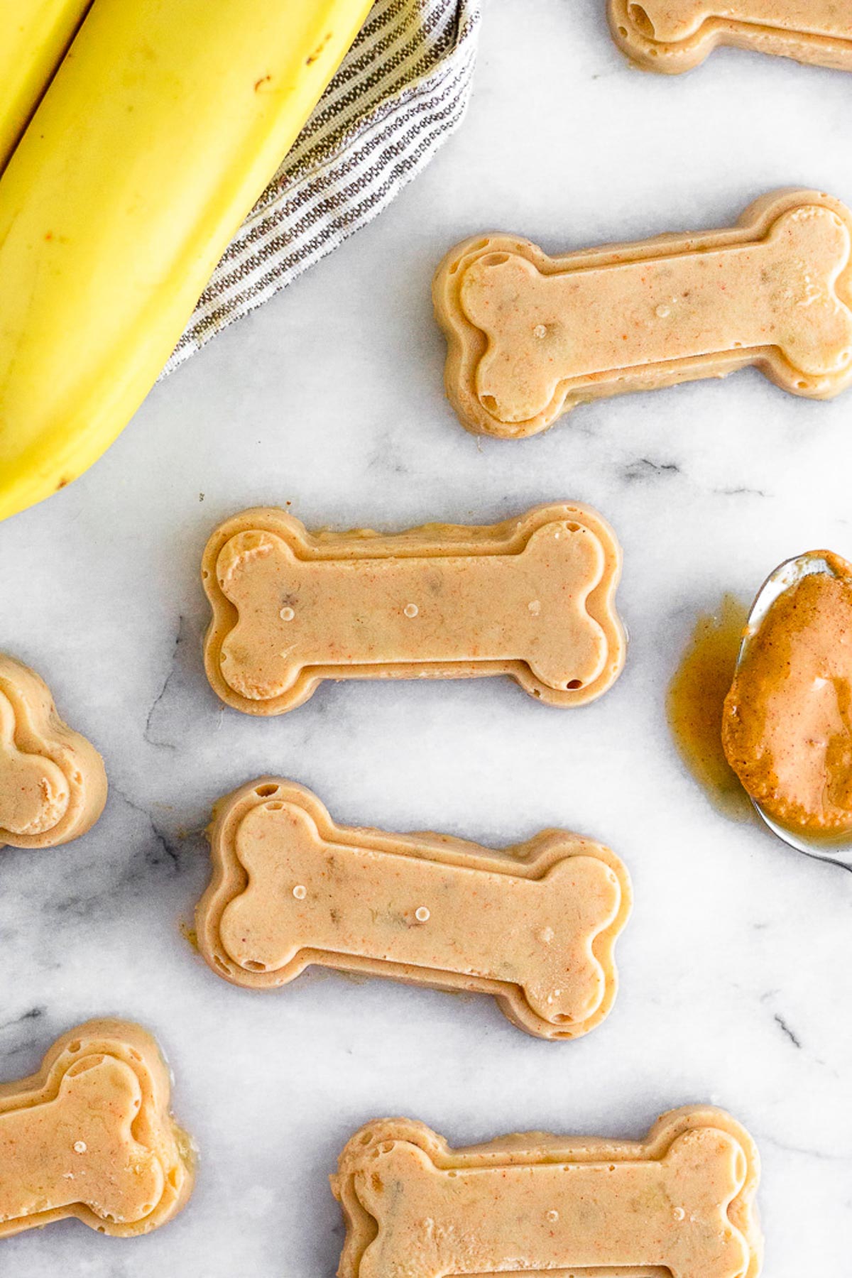 Overhead shot of peanut butter banana dog treats shaped as bones with a spoonful of peanut butter and bananas next to them.