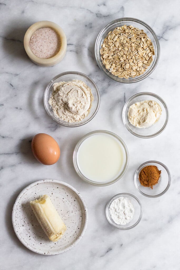 Marble counter with a bowl of rolled oats, a bowl of coconut flour, a small bowl of cinnamon, a small bowl of baking powder, a small plate with a half of banana, a brown egg, a bowl of milk, a bowl of protein powder, and a dish of salt.