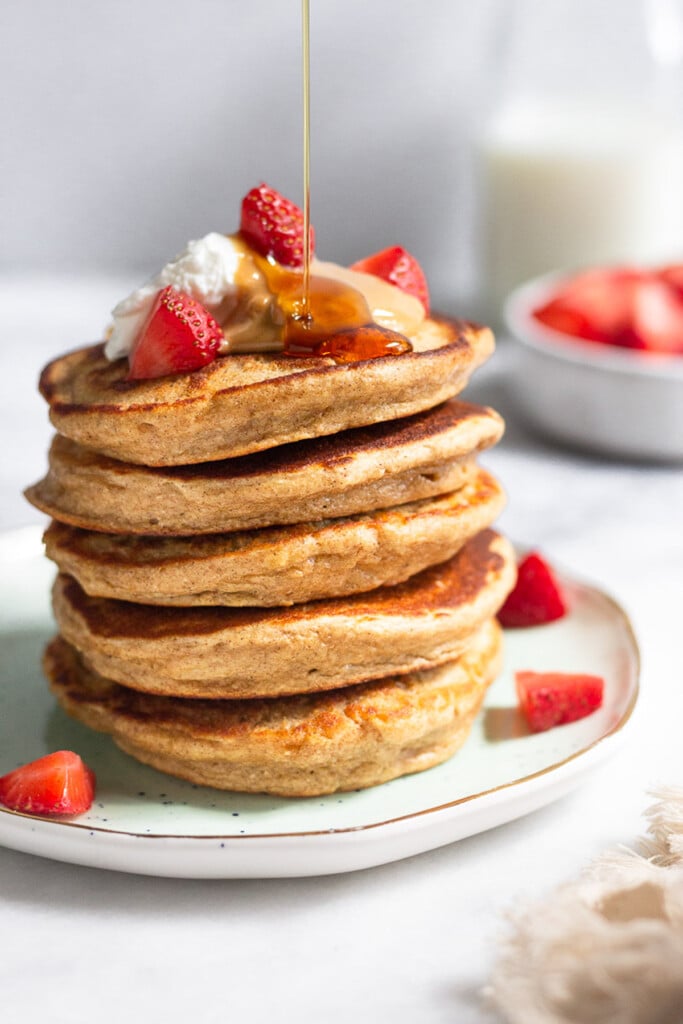 Plate with a stack of protein powder pancakes topped with strawberries, peanut butter, and yogurt with maple syrup being poured on them. Behind the plate is a bowl of diced strawberries and a jar of milk.