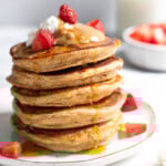 A stack of protein pancakes on a plate topped with yogurt, peanut butter, and strawberries with maple syrup dripping down them. Behind the plate is a bowl of diced strawberries and a jar of milk.