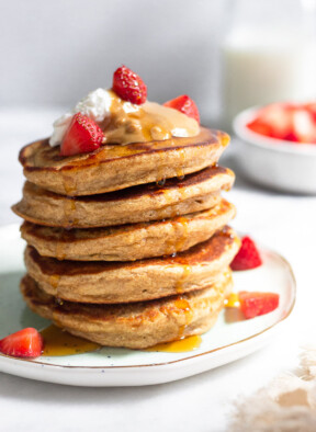 A stack of protein pancakes on a plate topped with yogurt, peanut butter, and strawberries with maple syrup dripping down them. Behind the plate is a bowl of diced strawberries and a jar of milk.