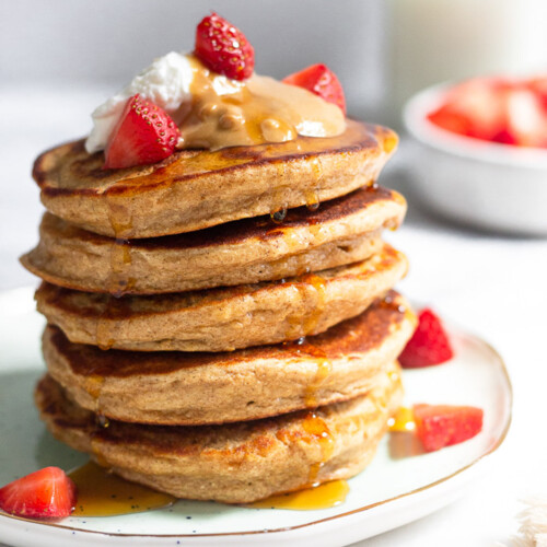 A stack of protein pancakes on a plate topped with yogurt, peanut butter, and strawberries with maple syrup dripping down them. Behind the plate is a bowl of diced strawberries and a jar of milk.