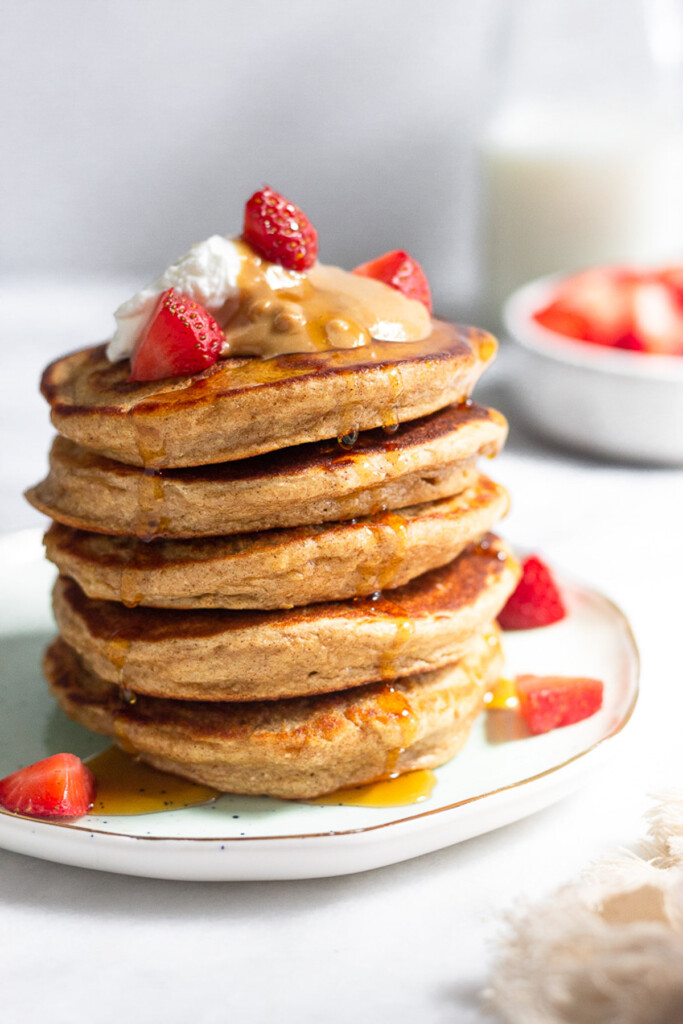 A stack of protein pancakes on a plate topped with yogurt, peanut butter, and strawberries with maple syrup dripping down them. Behind the plate is a bowl of diced strawberries and a jar of milk.