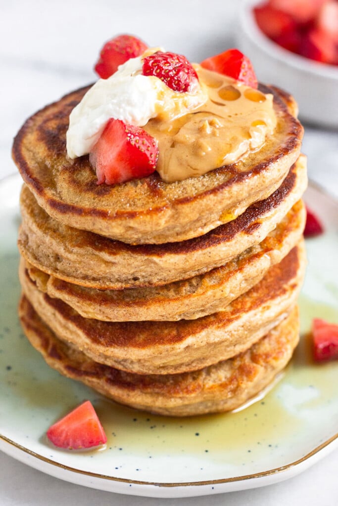 Close up of a large stack of gluten free protein pancakes topped with yogurt, peanut butter, and strawberries. Maple syrup has been poured overtop of them. Behind the plate of pancakes is a bowl of strawberries.