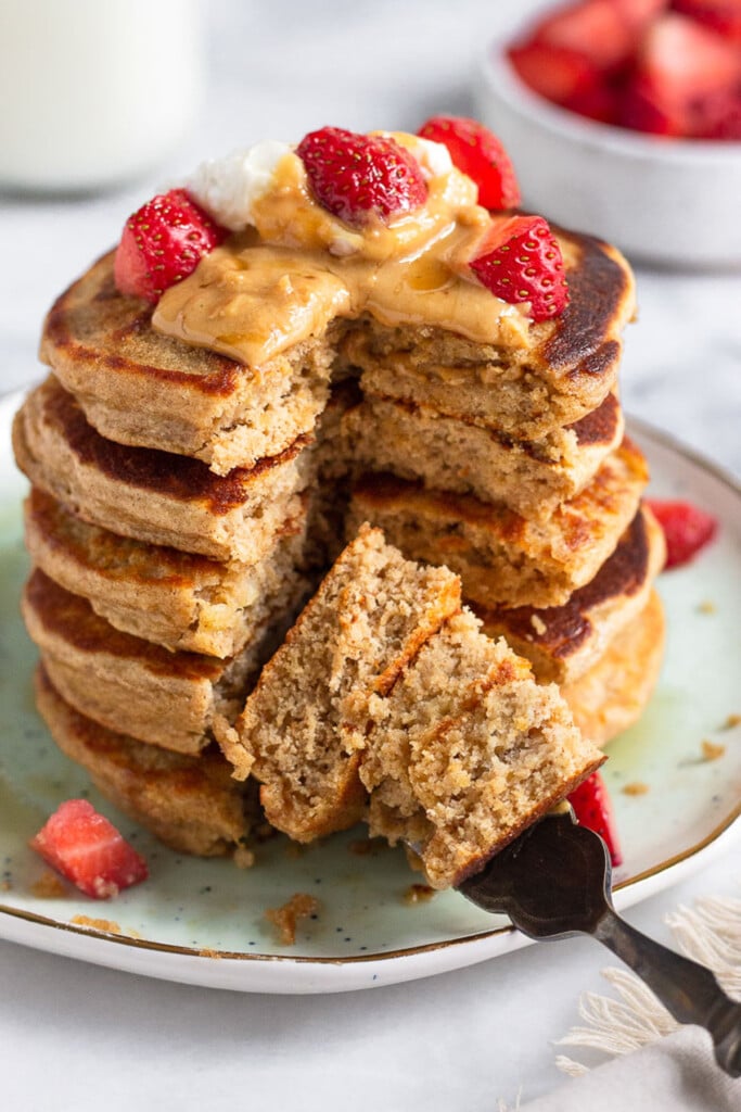 High protein pancakes stacked on a plate with a section cut out of them. They are topped with yogurt, peanut butter, and strawberries. There is a fork holding up some of the cut pieces in front of the plate. Behind the plate is a bowl of strawberries and a jar of milk.